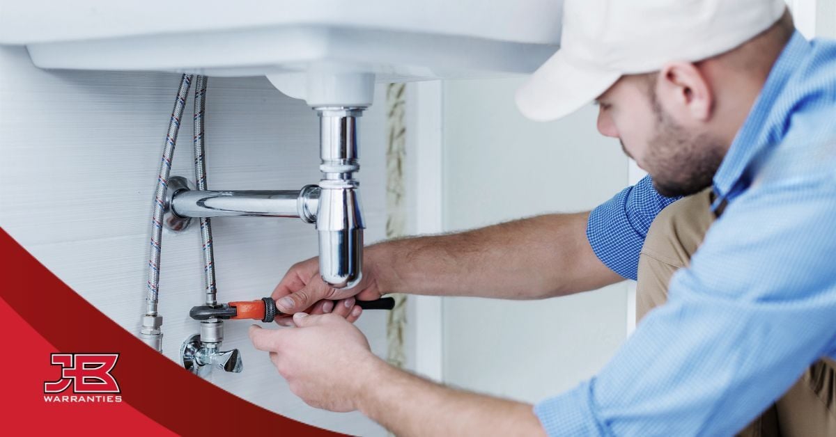 Plumber tightening a shutoff valve beneath a bathroom sink using a wrench while inspecting exposed plumbing connections. The image includes the JB Warranties logo in the lower-left corner.